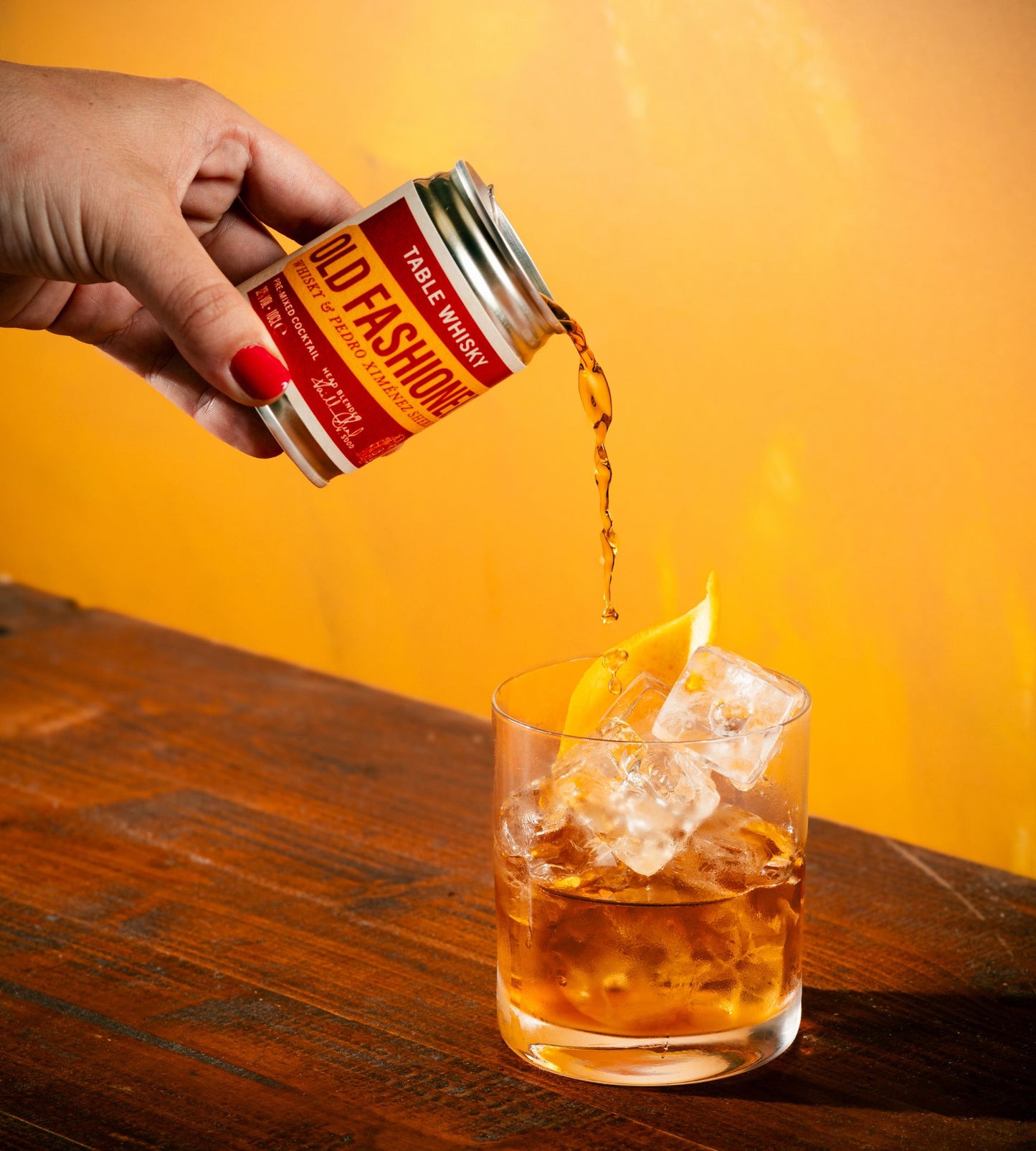 A hand with red nail polish pours a stream of amber liquid from a can of Old Fashioned Table Whiskey into a rocks glass filled with ice and garnished with an orange peel, set against a warm yellow background and dark wooden table.