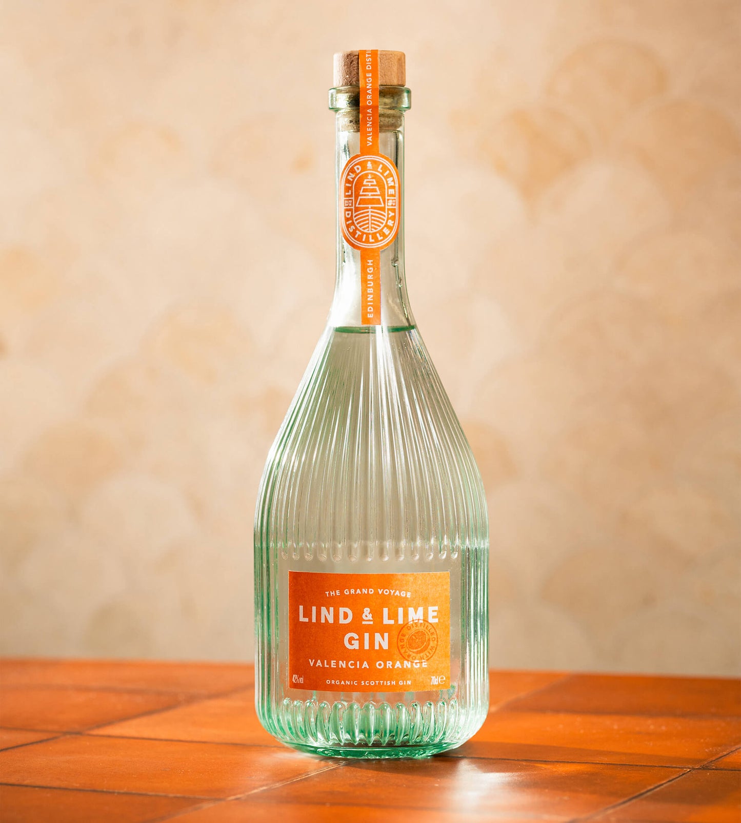 A bottle of Lind & Lime Gin Valencia Orange featuring a ribbed clear glass bottle with a vibrant orange label and neck seal, set on a terracotta tiled surface against a textured beige wall.