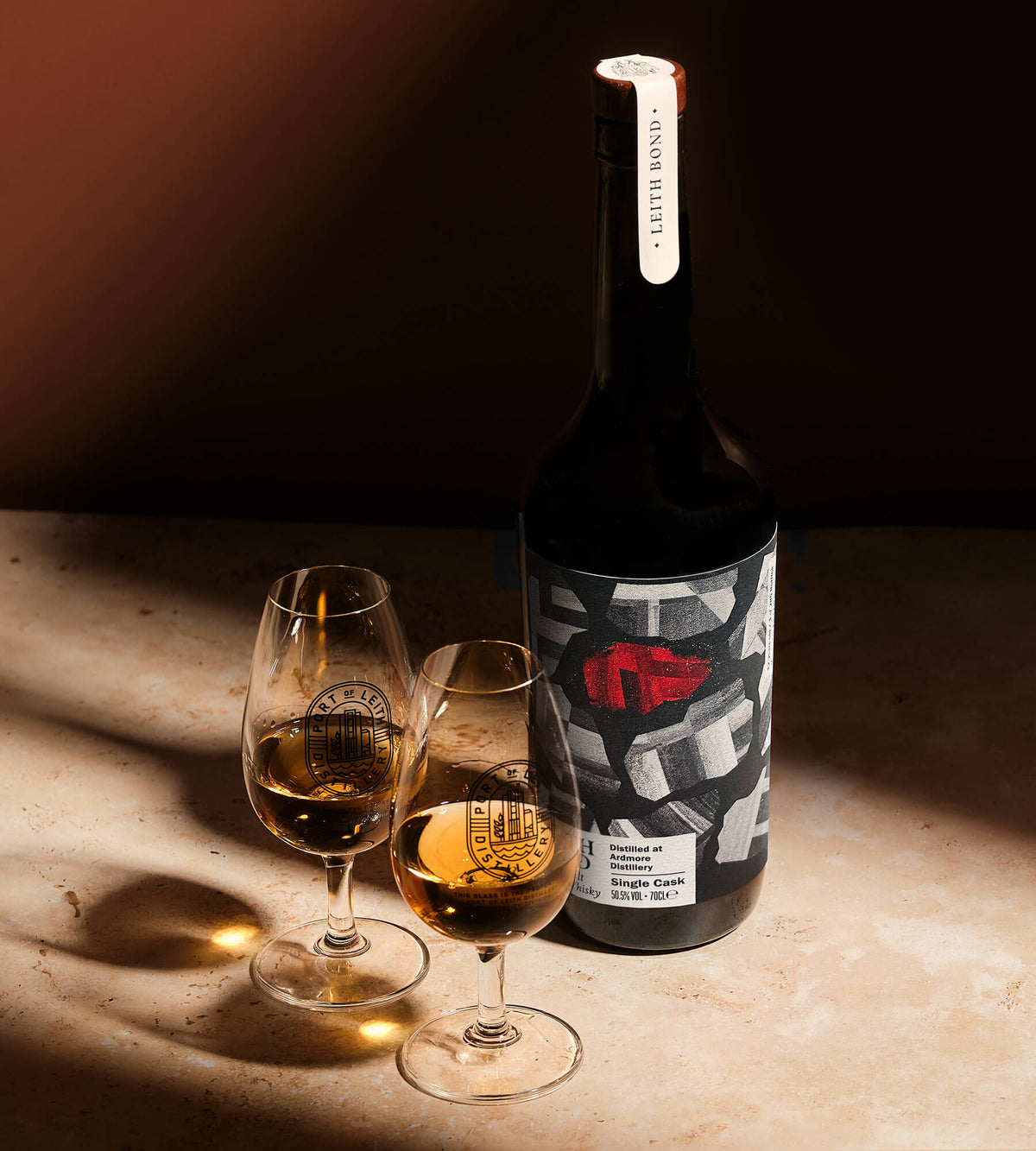 A dark whisky bottle with a graphic label featuring a red abstract design, standing next to two stemmed glasses filled with an amber-colored whisky on a warm, sunlit stone surface.