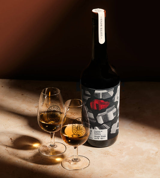 A dark whisky bottle with a graphic label featuring a red abstract design, standing next to two stemmed glasses filled with an amber-colored whisky on a warm, sunlit stone surface.