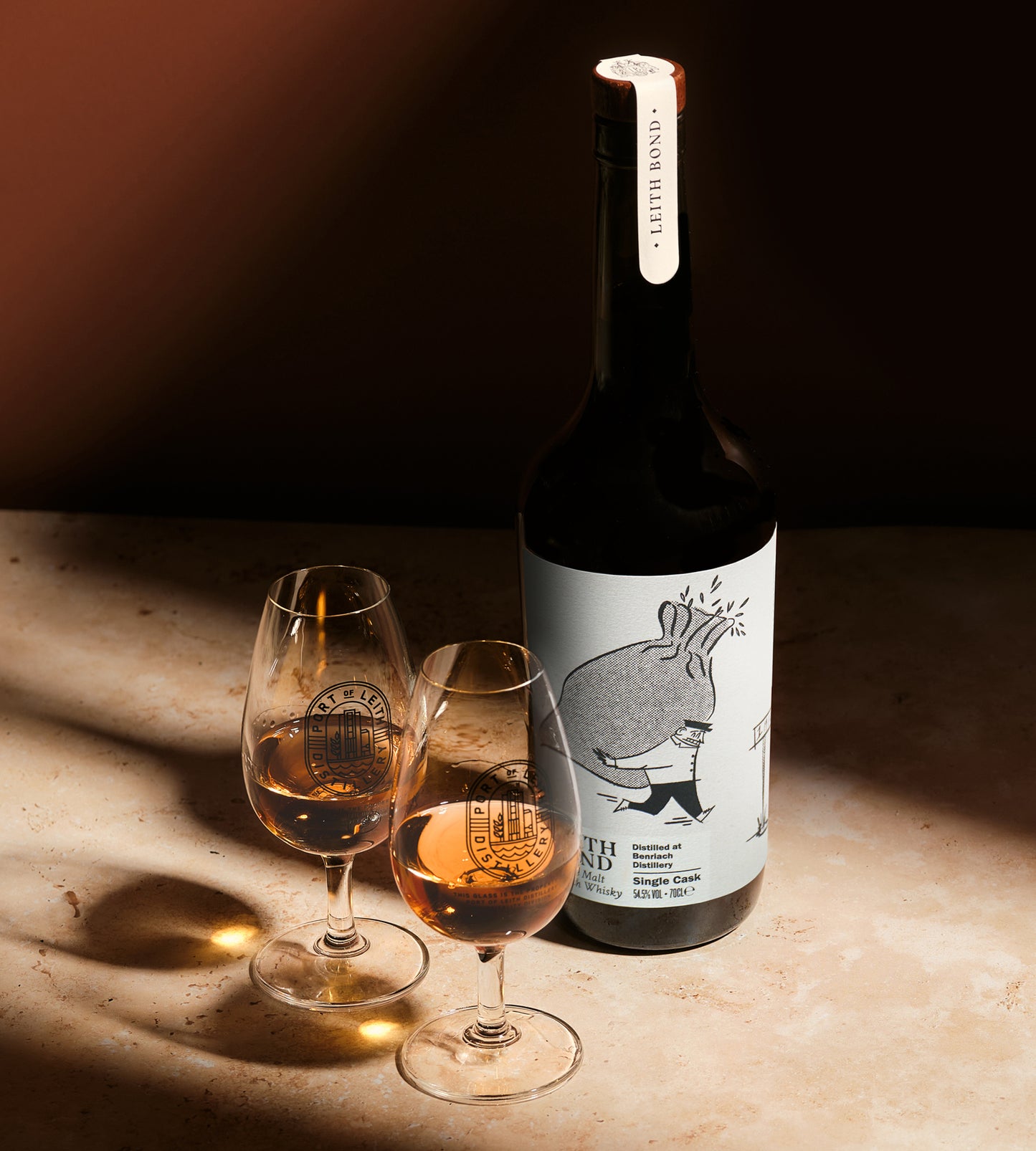 A lifestyle image of the Benriach 12yo Single Cask Whisky. The bottle is standing on a stone surface beside two nosing glasses filled with the whisky. The scene is lit by dramatic directional sunlight casting strong shadows.