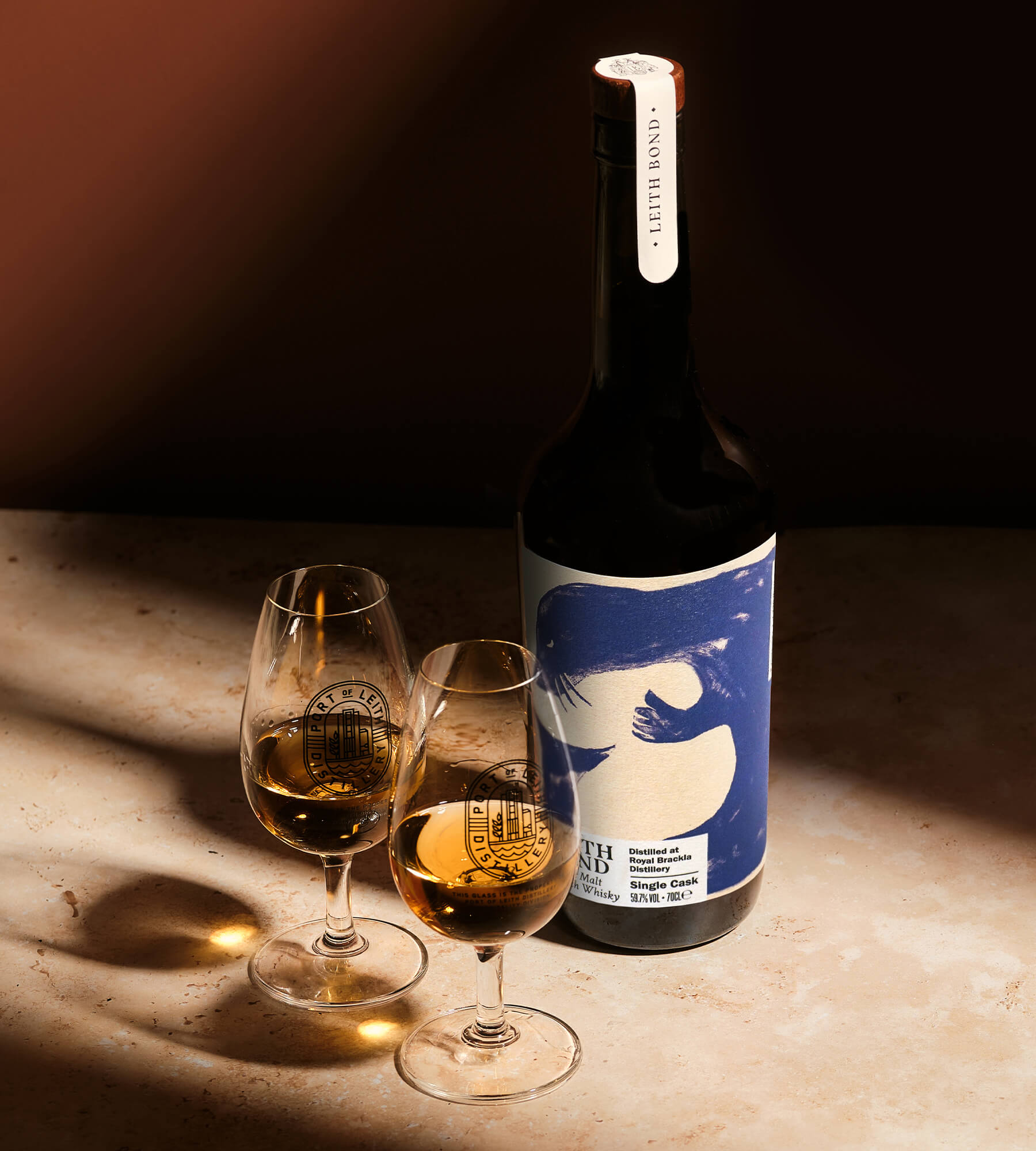 A bottle of Cawdor Springs Single Cask Whisky, distilled at Royal Brackla. Featuring a blue and cream graphic label, displayed next to two nosing glasses filled with whisky on a warm, sunlit stone surface.