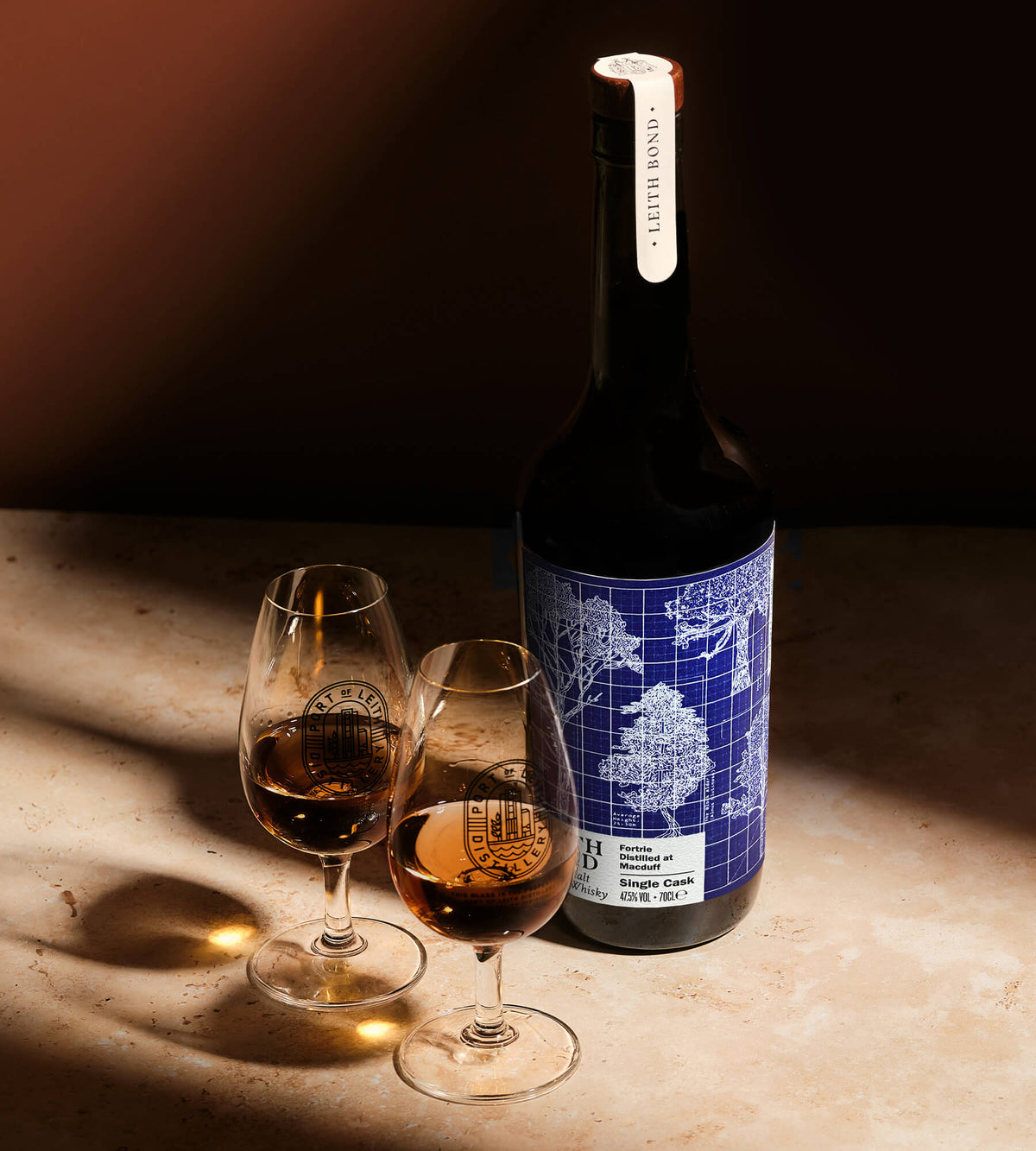 A bottle of single malt whisky with a blue and white architectural label stands on a stone surface beside two stemmed glasses filled with the wine, lit by dramatic directional sunlight.