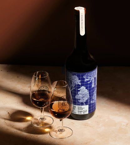 A bottle of single malt whisky with a blue and white architectural label stands on a stone surface beside two stemmed glasses filled with the wine, lit by dramatic directional sunlight.