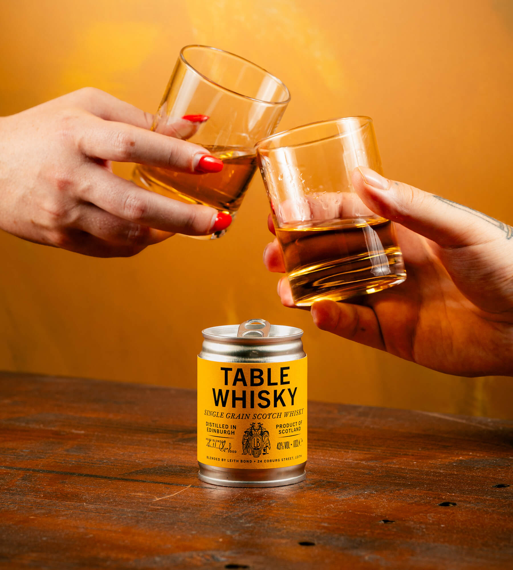 Two hands holding up clear rocks glasses filled with amber whisky above a small 100 ml can of Table Whisky on a wooden table, against a warm orange background.