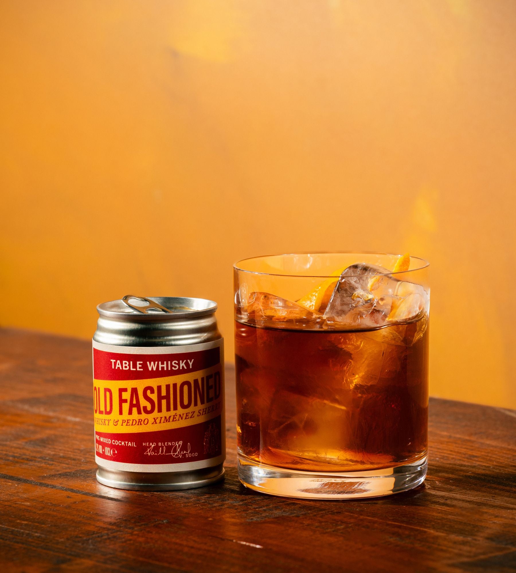 A small silver can of Table Whisky 'Old Fashion' table whisky next to a clear glass filled with an amber-brown drink and ice, set on a wooden table against a warm orange background.