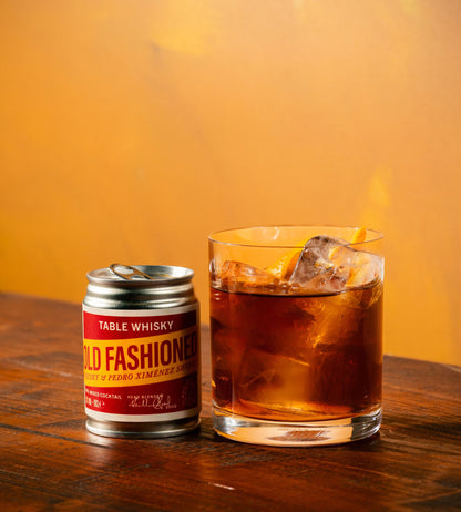 A small silver can of Table Whisky 'Old Fashion' table whisky next to a clear glass filled with an amber-brown drink and ice, set on a wooden table against a warm orange background.