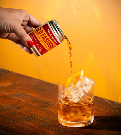 A hand with red nail polish pours a stream of amber liquid from a can of Old Fashioned Table Whiskey into a rocks glass filled with ice and garnished with an orange peel, set against a warm yellow background and dark wooden table.