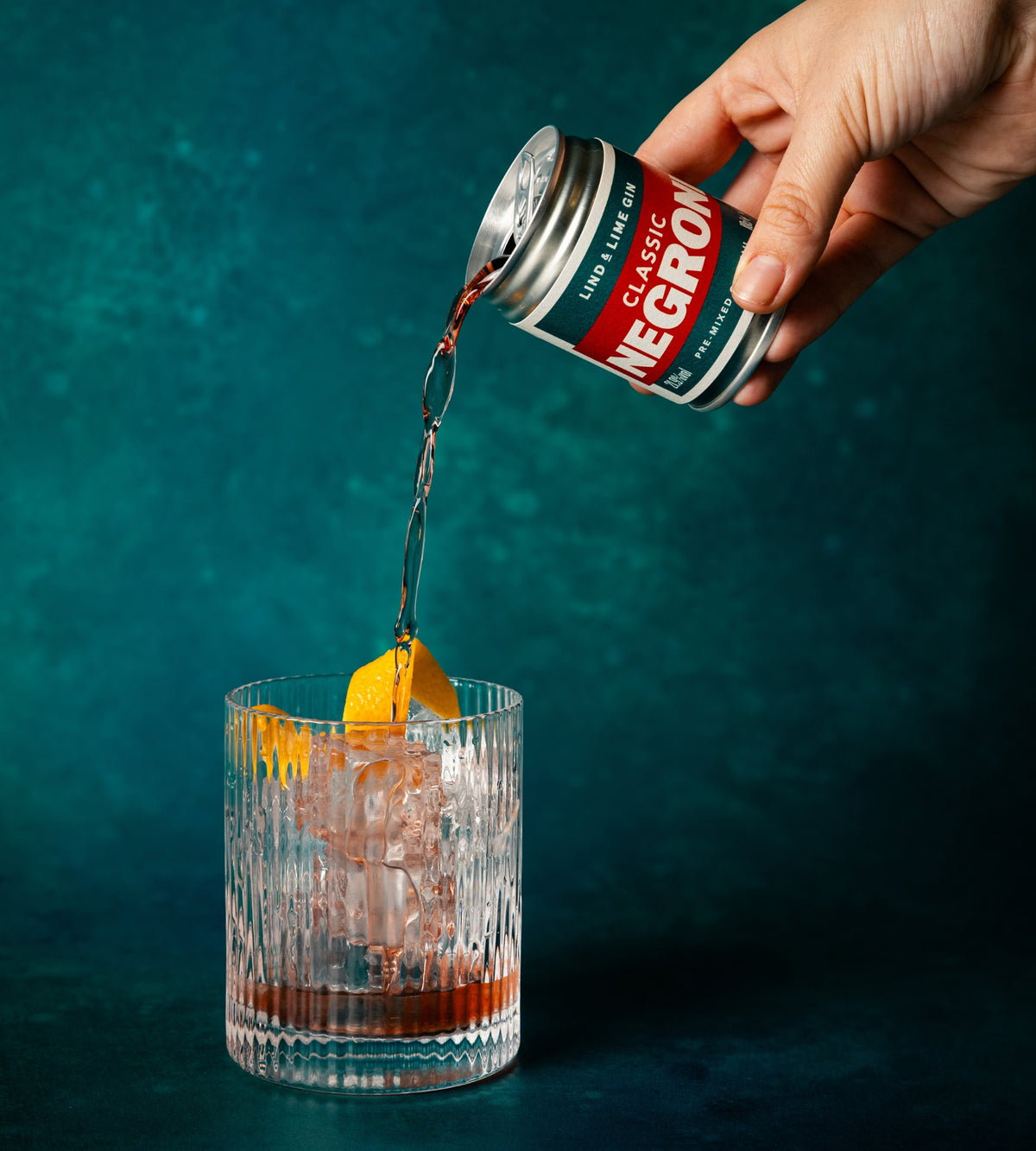 A hand pours Classic Negroni from a silver can into a clear textured rocks glass with ice and an orange peel garnish, set against a dark teal background.