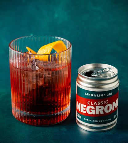 A glass of Negroni with ice and a orange peel garnish sits next to a can of Lind & Lime Gin Classic Negroni on a dark teal background.