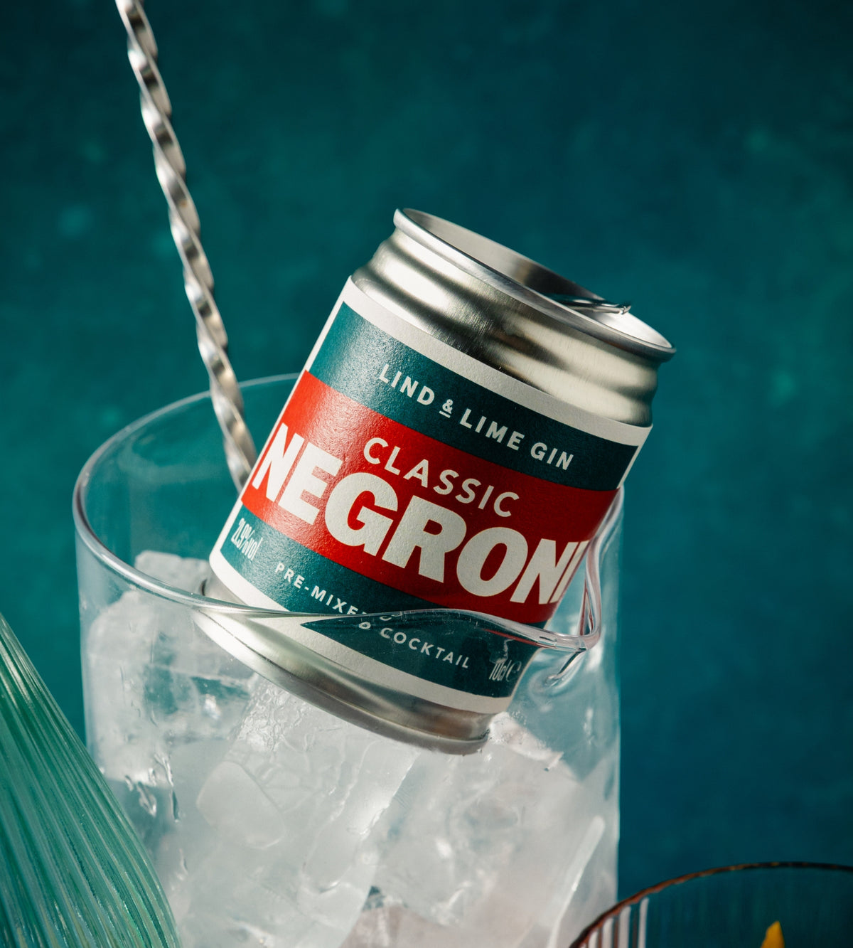 A can of Negroni pre-mix cocktail gin resting in a glass of ice, with a metal bar spoon, on a dark teal background.
