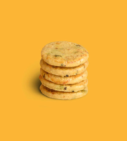 A stack of five round golden baked crackers or biscuits with visible green and dark flecks, centered on a solid yellow background.